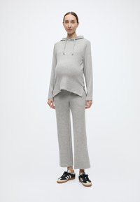 Pelēka grūtnieču kapuce ar kengura kabatu un auklu, kopā ar atbilstošām pelēkām plaša piegriezuma biksēm un melnām sporta kurpēm ar baltām detaļām.