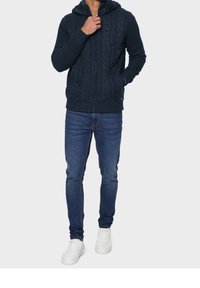 Marinblå kabelstickad hoodie med frontdragkedja, ribbade ärmslut och nederkant, i kombination med blå jeans och vita sneakers. Enkel och figursydd design.