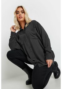 Camisa negra de gran tamaño hecha de tela suave, con botones en la parte delantera, dos bolsillos en el pecho y mangas relajadas. Combinada con pantalones negros y botas.