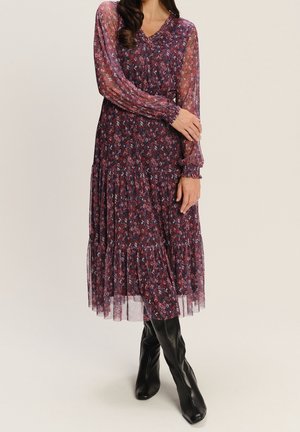 Frau trägt ein langärmeliges, dunkles florales Midikleid mit transparenten Ärmeln, kombiniert mit schwarzen kniehohen Stiefeln, steht mit verschränkten Armen.