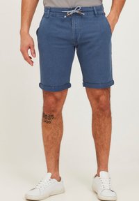 Blue cotton shorts with a drawstring detailing, rolled cuffs, and side pockets, paired with white sneakers. Features a visible tattoo.
