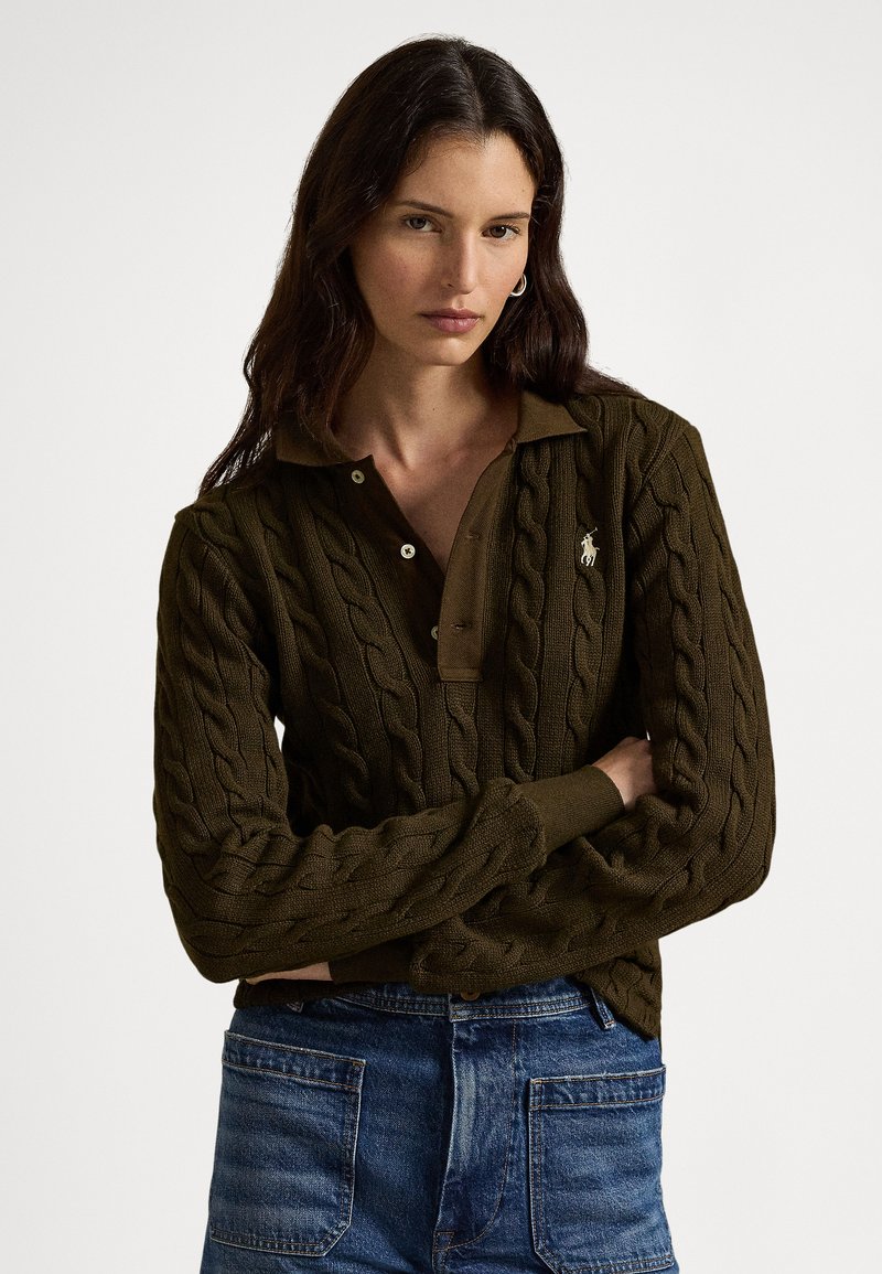 Γυναίκα με μακριά σκούρα μαλλιά, φοράει σκούρο πράσινο πουλόβερ πλεκτό με γιακά και μπλε τζιν, στέκεται με τα χέρια σταυρωμένα μπροστά σε λευκό φόντο.