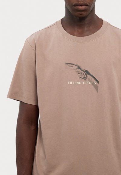Beige Baumwoll-T-Shirt mit kurzen Ärmeln, das eine Grafik von zwei Händen und den Text "Filling Pieces" in Weiß zeigt. Einfaches, lässiges Design.
