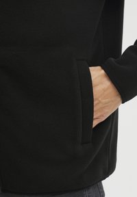 Hånd indsætter sig i for lommen på en sort fleecejakke, der bæres over mørke jeans, som viser stoftekstur og kant på ærmet.