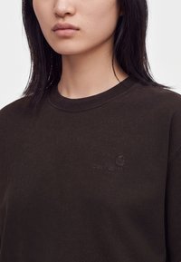 Γυναίκα που φοράει ένα σκούρο καφέ φούτερ Carhartt με λογότυπο στο στήθος, εμφανιζόμενη από τον λαιμό μέχρι το μέσο του κορμού σε απλό φόντο.