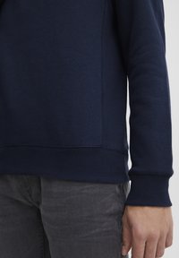 Navy blue sweatshirt with ribbed cuffs and hem, featuring a smooth texture and visible seam details. Paired with gray jeans.