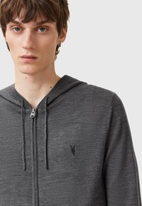 Sweat à capuche gris à fermeture éclair avec une finition texturée, doté d'une capuche, de cordons de serrage et d'un petit logo noir sur la poitrine. La fermeture éclair est visible.