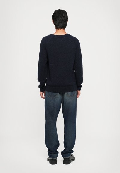 Pulôver texturizado azul-marinho com decote redondo, mangas longas e punhos canelados, combinado com jeans azuis largos e sapatos pretos.