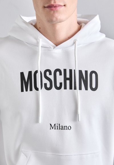 Hanorac alb din bumbac, cu text îndrăzneț negru "MOSCHINO" și text mai mic "Milano". Include șnur de ajustare și buzunar frontal tip kangaroo.