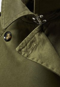Vihreä oliivinvärinen trench coat -takki, jossa on suuri kaulus, kiiltävä musta nappi ja hopeiset yksityiskohdat kiinnityksessä. Suuri kangas on sileäpintainen.