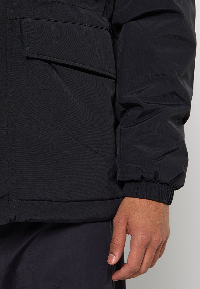 Giacca isolante nera con superficie testurizzata, polsini elasticizzati e tasche laterali. Presenta una vestibilità morbida e un design minimalista.