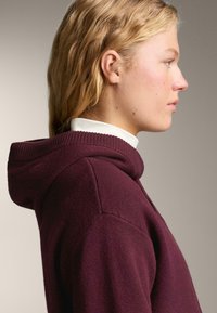 Blondínka v profilu, ktorá má na sebe bordový mikinu nad bielym rolákom na neutrálnom pozadí.