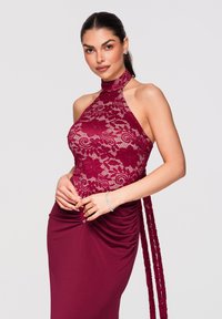 Femme portant une robe bordeaux sans manches en dentelle avec un motif floral, jupe froncée, bracelet argenté et boucles d'oreilles, posant les mains jointes.