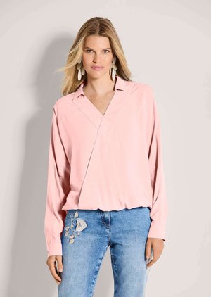 Blonde Frau trägt eine übergroße hellrosa Bluse und hellblaue Jeans mit Blumenstickerei und steht vor einem hellgrauen Hintergrund.