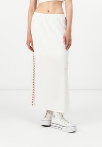 Falda maxi blanca con una cinturilla elástica, que presenta detalles a cuadros naranjas en las costuras laterales. Combinada con zapatillas altas blancas.