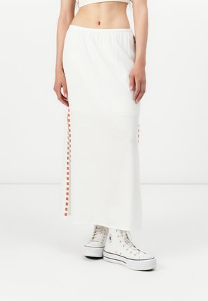 Falda maxi blanca con una cinturilla elástica, que presenta detalles a cuadros naranjas en las costuras laterales. Combinada con zapatillas altas blancas.