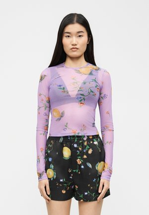 Jauna moteris su ilgais tamsiais plaukais dėvi permatomą levandų spalvos gėlių rašto viršutinę dalį ir juodus gėlių rašto šortus, stovinčią priešais paprastą baltą foną.