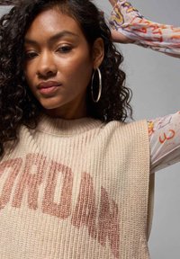 Beige ribbestrikket genser med en stor "JORDAN" logo i falmet rosa. Langermet mønstret topp synlig under. Sølv hoopsøreringer.