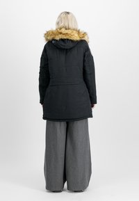 Parka isolante noire avec capuche bordée de fourrure, manches longues et taille ajustée. Portée sur un pantalon gris à jambes larges, design à fort contraste.