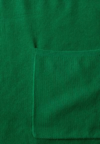 Grön stickad tyg med en texturerad yta och synlig fickdetalj. Tyget är jämnt sytt med en ribbad nederkant.
