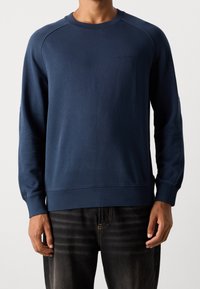 Uomo che indossa un semplice felpa a maniche lunghe blu navy e jeans neri, in piedi contro uno sfondo bianco.