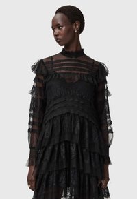 Svart spetsklänning med lager av volanger, genomskinliga ärmar och hög krage. Strukturera mönster genom hela klänningen med en figursydd bodice.