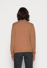 Osoba z włosami do ramion w ciemnym kolorze, ubrana w brązowy sweter z długim rękawem i czarne dżinsy, zwrócona plecami w stosunku do jednolitego tła.