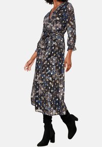 Žena v černých květinových šatech s dlouhým rukávem a černých středně vysokých botách na podpatku, kráčející na bílém pozadí.