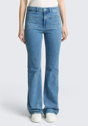 Femme portant un jean droit taille haute bleu clair avec des poches appliquées à l'avant et des baskets blanches, debout devant un fond uni.