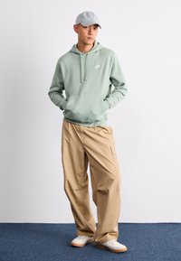 Fleece verde claro com bolso frontal, calças cargo bege e boné cinza. Ténis brancos com sola de borracha. Fundo simples e chão azul.