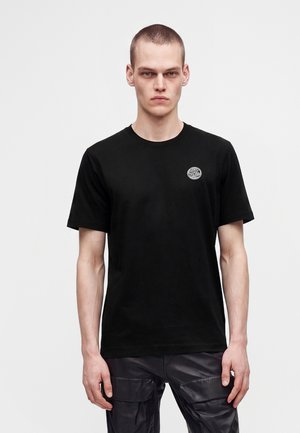 Joven con el pelo corto que lleva una camiseta negra con un pequeño logo redondo en el pecho y pantalones cargo negros, de pie frente a un fondo liso.