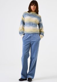 Vrouw in een pastelgekleurde trui met golvende patronen en losse blauwe broek, staande met haar handen in de zakken tegen een effen witte achtergrond.