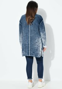 Langer Denim-Jacke in einem blauen Farbverlauf, weiche Textur, mit asymmetrischem Saum und weißen Nähten. Kombiniert mit tiefblauen Jeans und Sneakers.