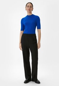 Blauw truitje met korte mouwen, gecombineerd met zwarte, loszittende, elastische broek. Het model draagt zwarte schoenen en zilveren oorringen.