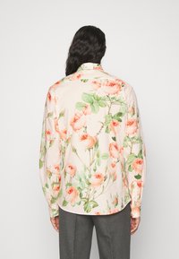 Persona con cabello largo y oscuro que lleva una camisa crema con un patrón floral de rosas rosas y hojas verdes, combinado con pantalones grises, vista por detrás.