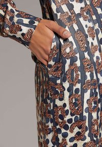 Kleid mit Leopardendruck, das aus einem strukturierten Stoff in Blau und Beige besteht, mit auffälligen dunklen Flecken und einem abstrakten Muster, ausgestattet mit seitlichen Taschen.