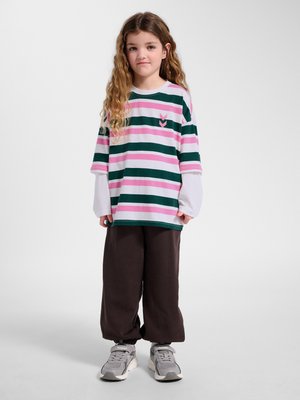 Jong meisje met lang krullend haar, die een oversized roze-groen gestreept overhemd draagt, bruine losse broek en grijze sneakers, staat stil.