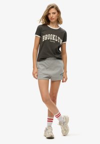 Camiseta gris con ribetes blancos y texto "BROOKLYN", combinada con pantalones cortos gris claro que tienen el texto azul "SUPERDRY NY". Calcetines blancos con franjas rojas.