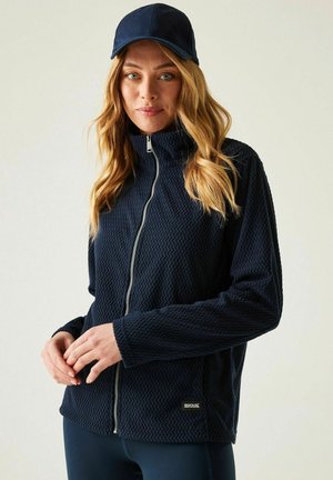 Kobieta w granatowej, teksturowanej kurtce na zamek i pasującej czapce, stojąca na jasnym, jednolitym tle, z rękami swobodnie złożonymi.