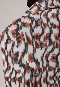 Veste ajustée en tissu texturé avec un motif ondulé dans des teintes orange, noir et blanc ; revers détaillé et épaules structurées.