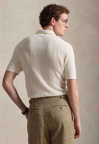 Άνδρας με σγουρά μαλλιά και γυαλιά, που φοράει μια υφασμάτινη εκρού κοντομάνικη μπλούζα και καφέ καρό ψηλόμεσο παντελόνι, στραμμένος πλευρικά.