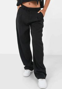 Zwarte, op maat gemaakte trousers met een losse pasvorm, met een hoge taille, plooien aan de voorkant en zijzakken. Gekleed met witte sneakers.