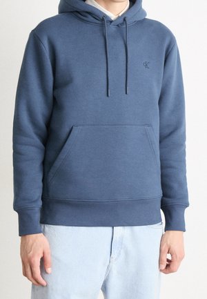 Osoba ubrana w niebieską bluzę Calvin Klein z kapturem i przednią kieszenią oraz jasnoniebieskie jeansy, stojąca na jednolitym białym tle.
