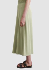 Light green, high-waisted maxi skirt made of soft fabric, with a flowy silhouette. Worn with black sandals featuring buckles.