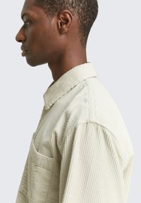 Beige ribfluweelenshirt met een kraag, zijzak en geribbelde textuur, getoond in profiel tegen een lichte achtergrond.