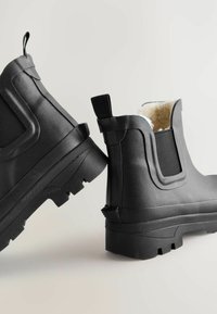 Bottes de cheville noires imperméables en matériau caoutchouteux texturé, avec des panneaux latéraux élastiques et une doublure douce. Semelle épaisse avec un profond relief.