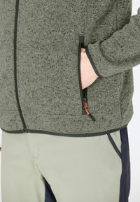 Gråspräcklig zip-jacka med ståkrage, som har två sidofickor, svart trim och en orange dragtab på dragkedjan.
