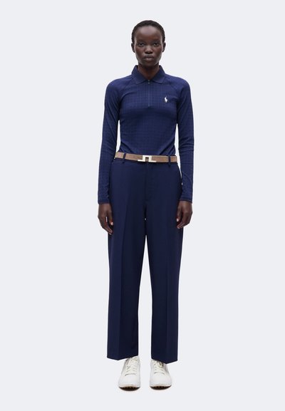 Femme portant un polo bleu marine à manches longues, un pantalon bleu marine assorti avec une ceinture marron, et des baskets blanches, debout devant un fond uni.