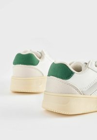Vita sneakers med gröna mockadetaljer, texturerad häl och gummisula. Har en snitsig design med en minimalistisk estetik.