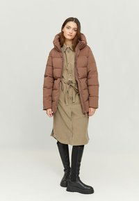 Brown puffer jacket with a hood, large quilted sections; worn over a beige button-up dress; paired with knee-high black boots.